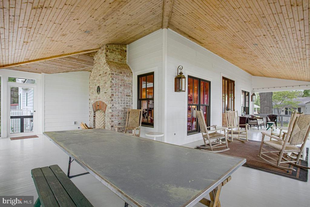 Covered porch with a brick fireplace, large table, and rocking chairs beneath a wood-plank ceiling.