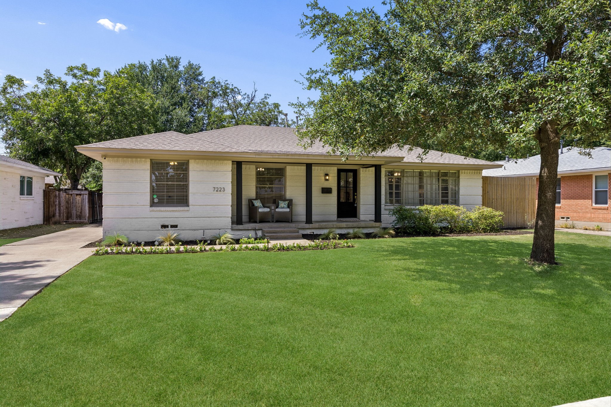 Single-story home with a covered porch, dark columns, a broad green lawn, and mature shade trees.
