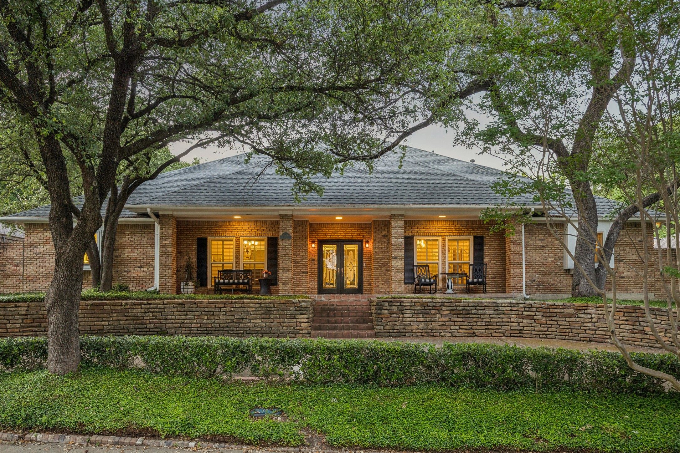 Brick home with decorative double doors, porch seating, and mature trees at dusk.