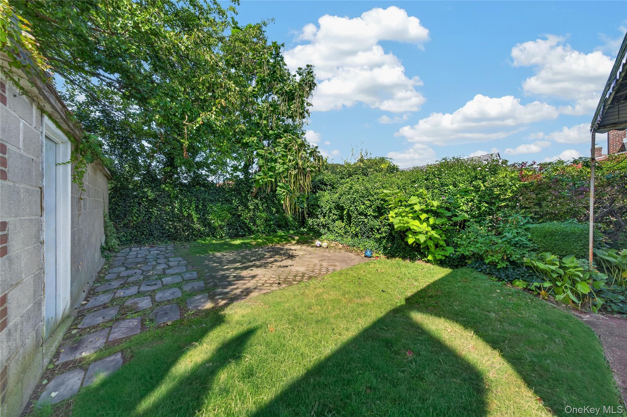 Enclosed backyard with a lawn, mature trees, tall hedges, and a stone-paver path.