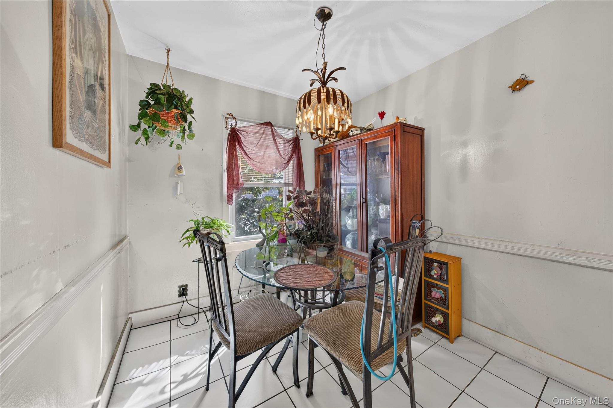 Dining room with a glass-top table, wooden china cabinet, hanging plants, and ornate chandelier.