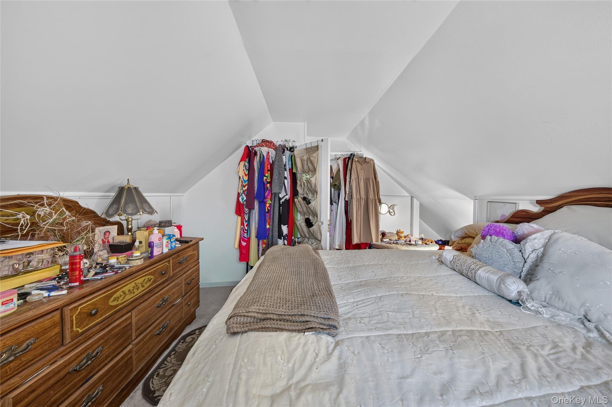 Attic bedroom with a large bed, sloped ceiling, open clothing racks, and a wooden dresser.