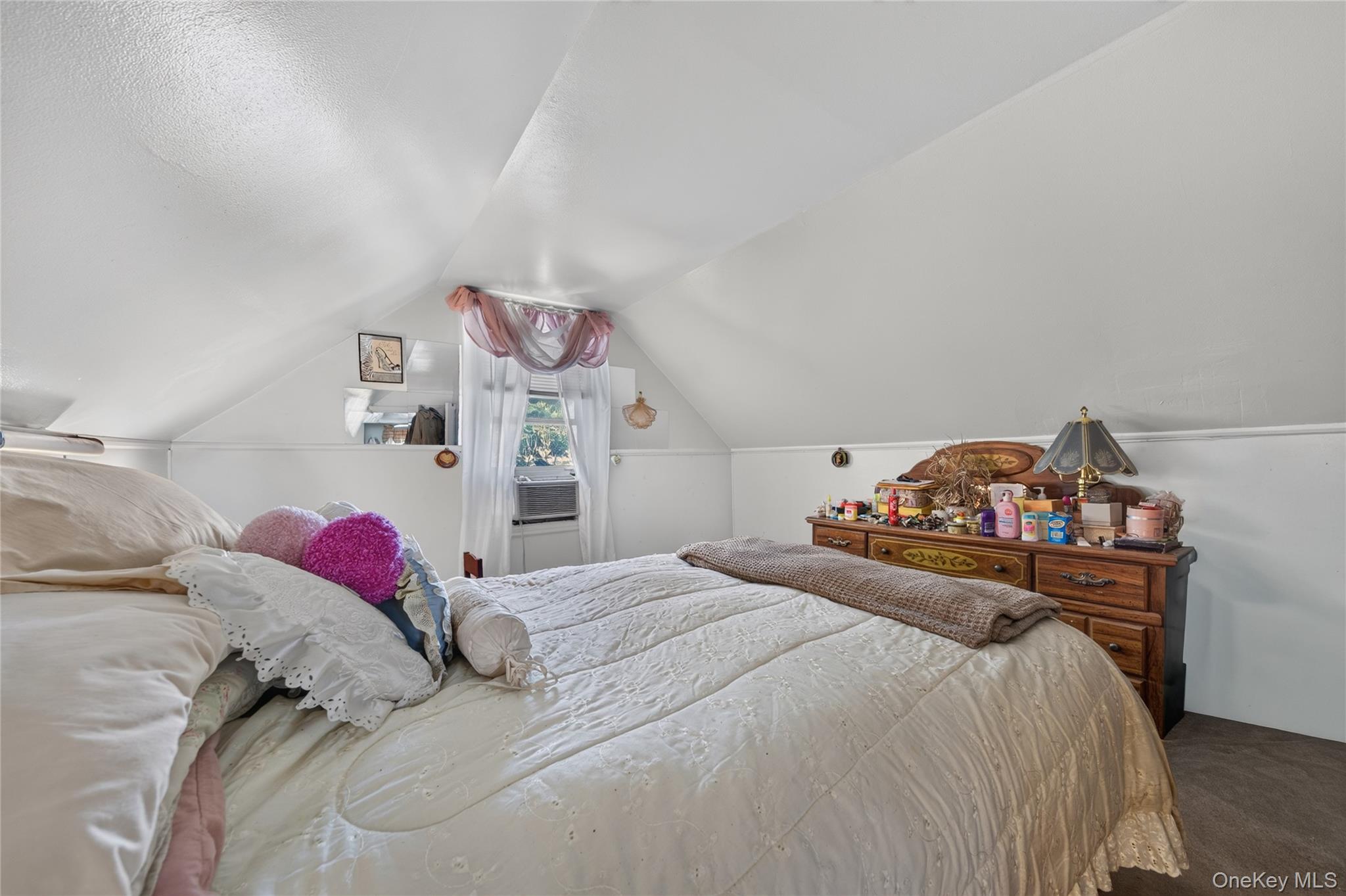 Attic bedroom with a large bed, sloped ceiling, curtained window, and wooden dresser.