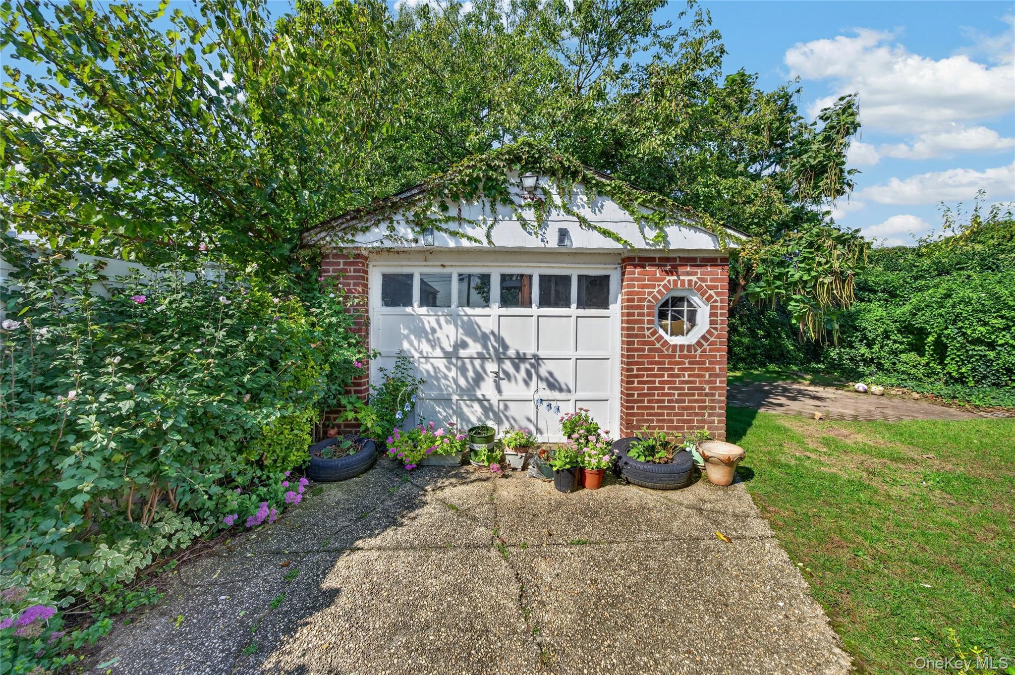 Brick garage with a white paneled door, octagonal window, climbing vines, and potted flowers.