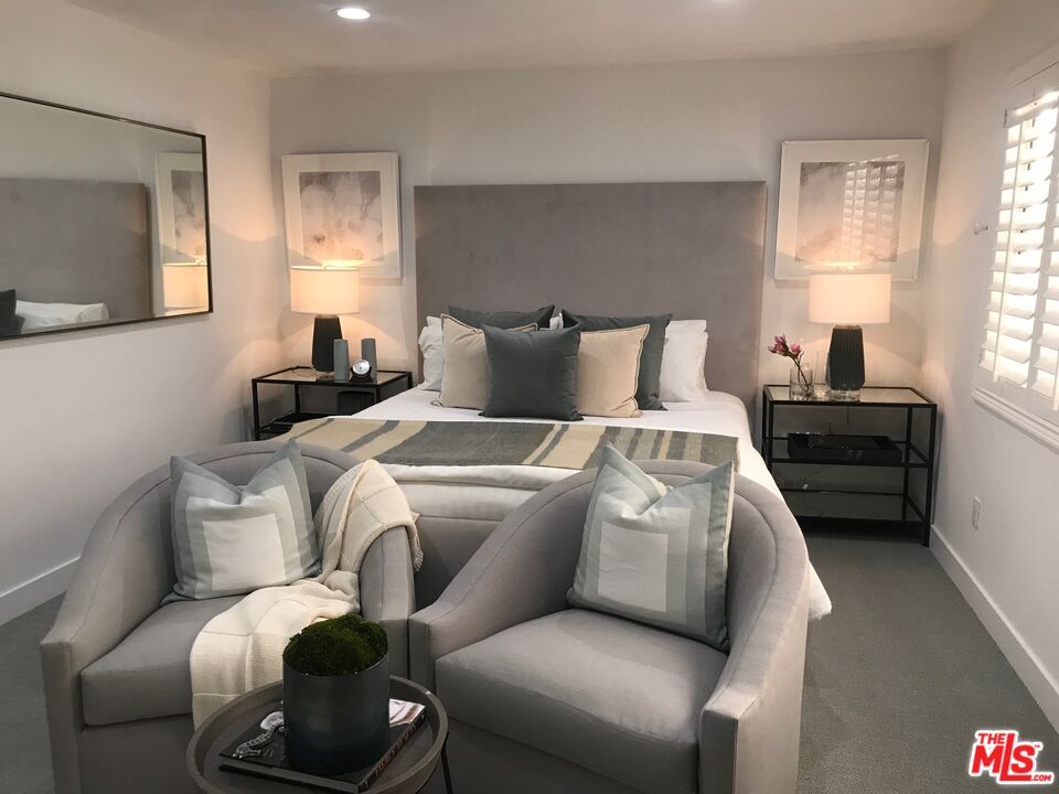 Staged bedroom with a large gray-headboard bed and two curved armchairs in the foreground.