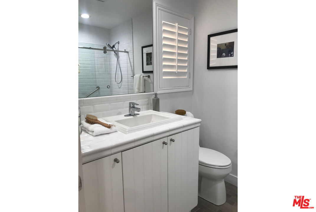White bathroom vanity and sink with a glass shower reflected in the mirror and a toilet beside it.