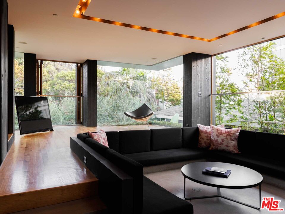 Modern living room with a black sectional sofa and expansive windows overlooking leafy gardens.