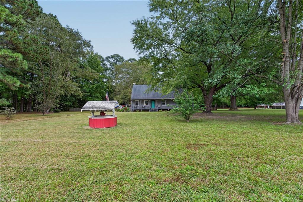 Gray single-story house with a teal door, broad lawn, mature trees, and a red wishing well.