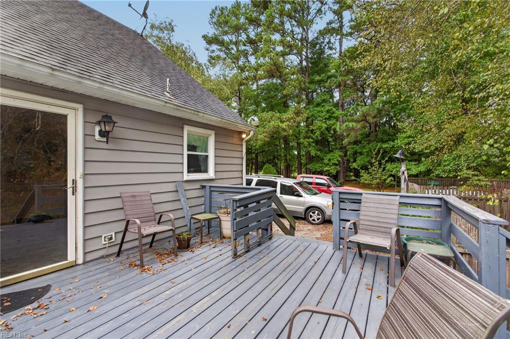 Gray-sided home with a spacious deck, outdoor chairs, parked cars, and a wooded yard.