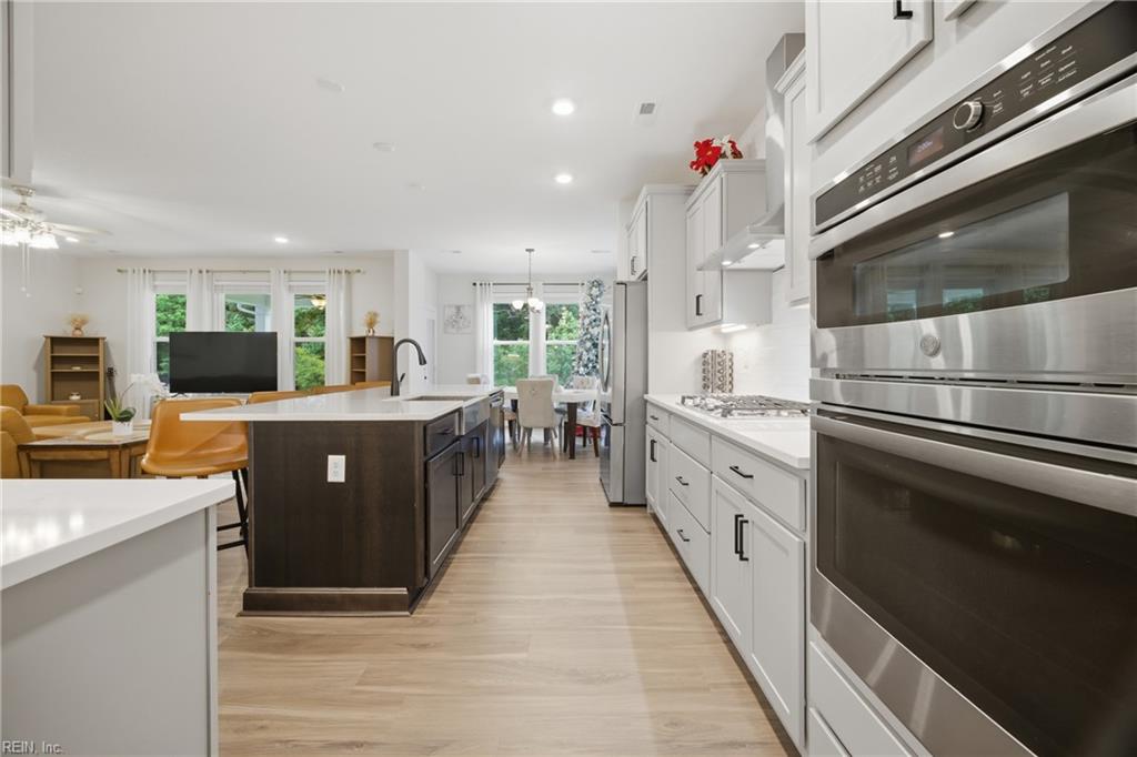 Open-plan kitchen with white cabinets, stainless steel double ovens, and a dark island overlooking the dining and living areas.