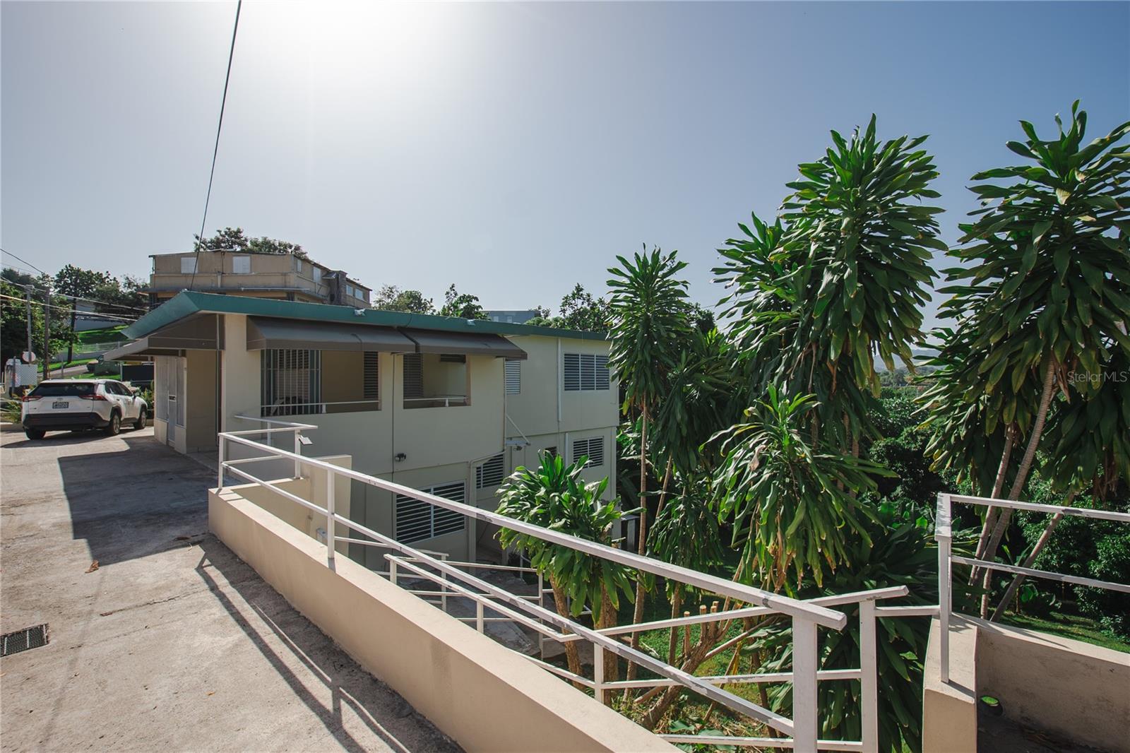 Light-colored multi-level building with a covered terrace, white railings, and tropical trees.