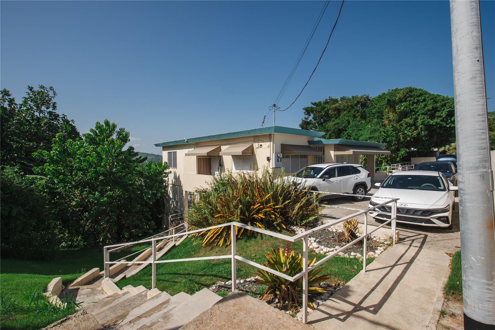 Low-rise cream building with a teal roof, parked cars, and a railed walkway amid tropical landscaping.