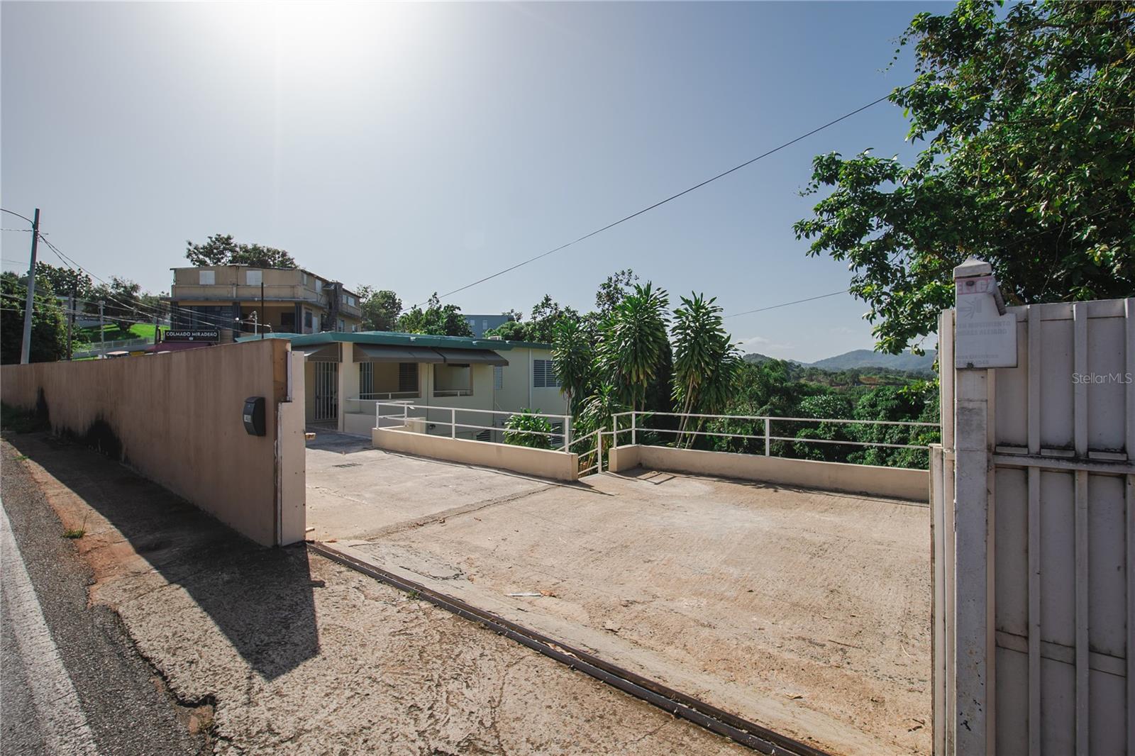 Low single-story property behind a concrete driveway and metal gate, with tropical greenery and distant hills.