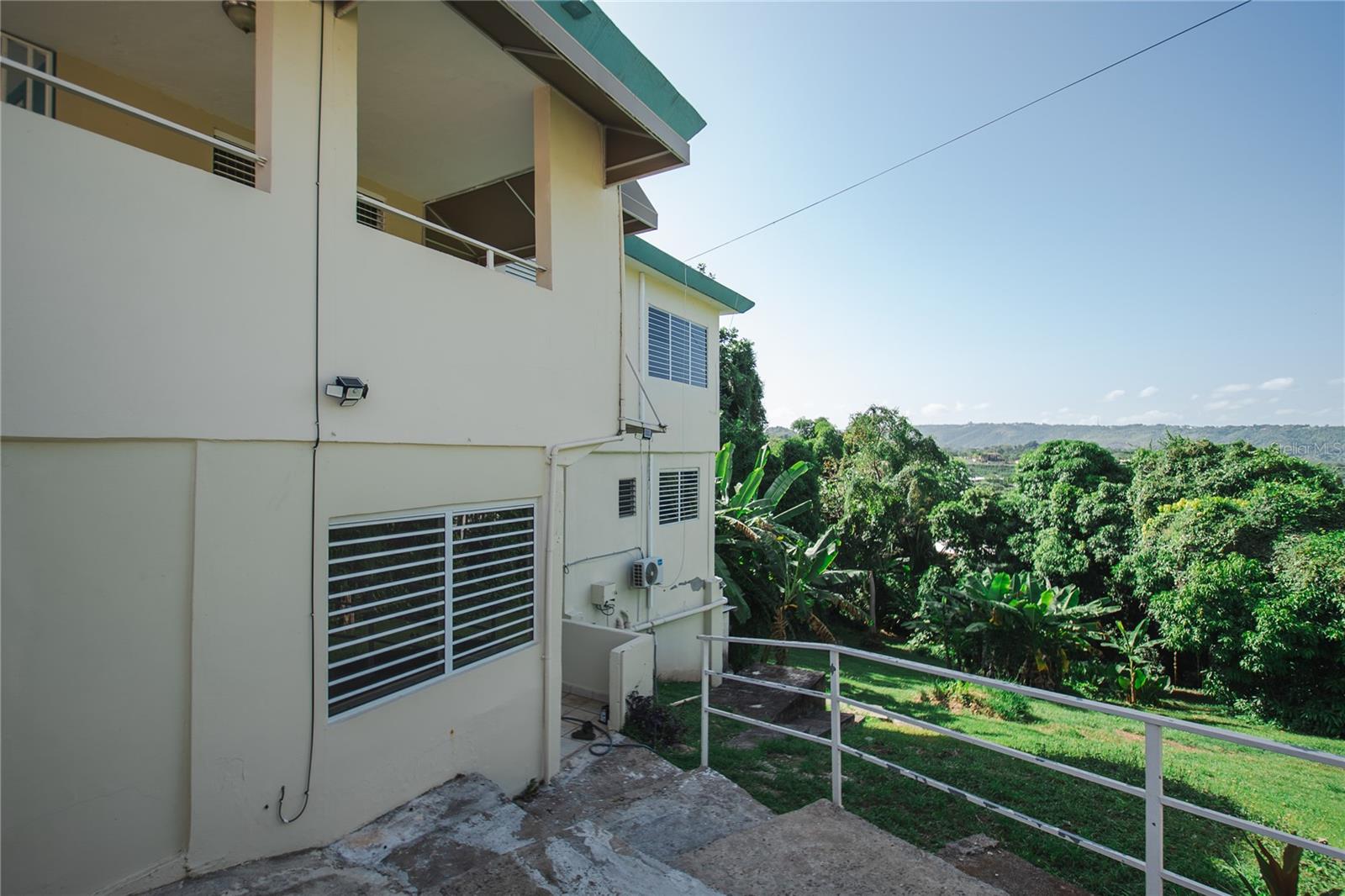 Cream-colored multi-level property beside a tropical yard and distant rolling hills.