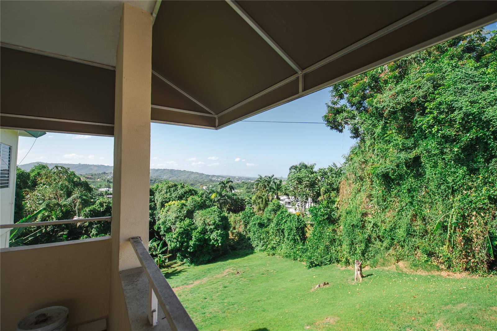 Covered balcony overlooking a green lawn, tropical trees, and distant rolling hills.