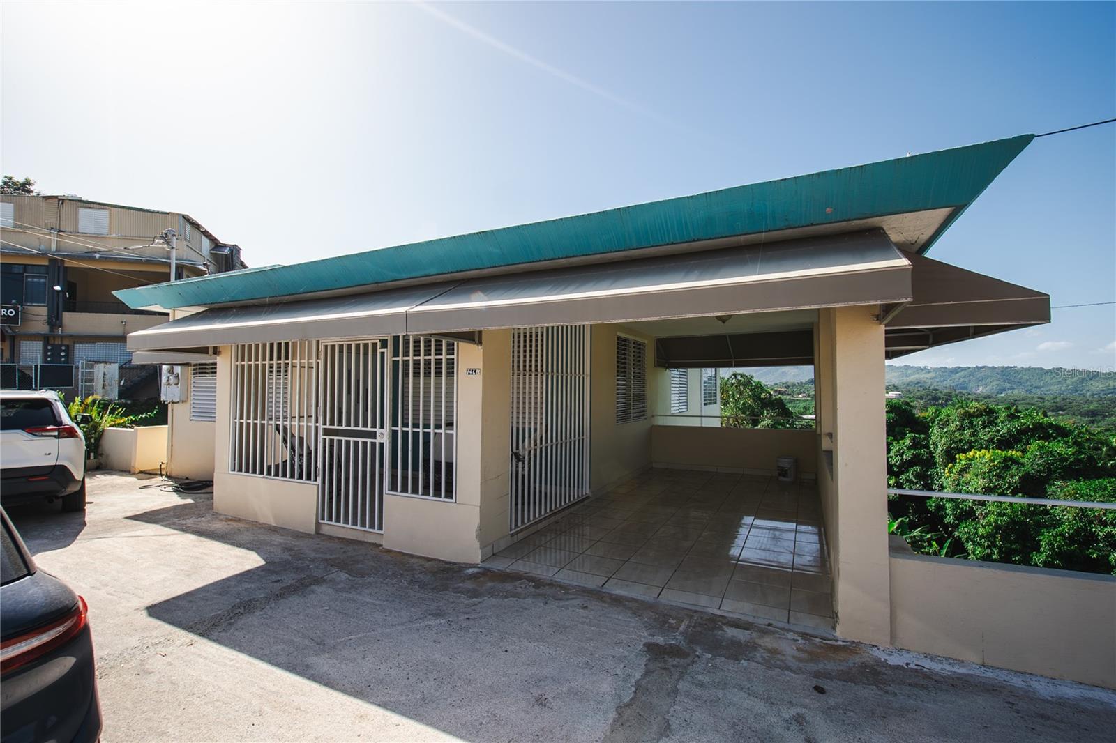 Beige home with barred windows, a tiled covered terrace, and a broad turquoise roof overlooking green hills.