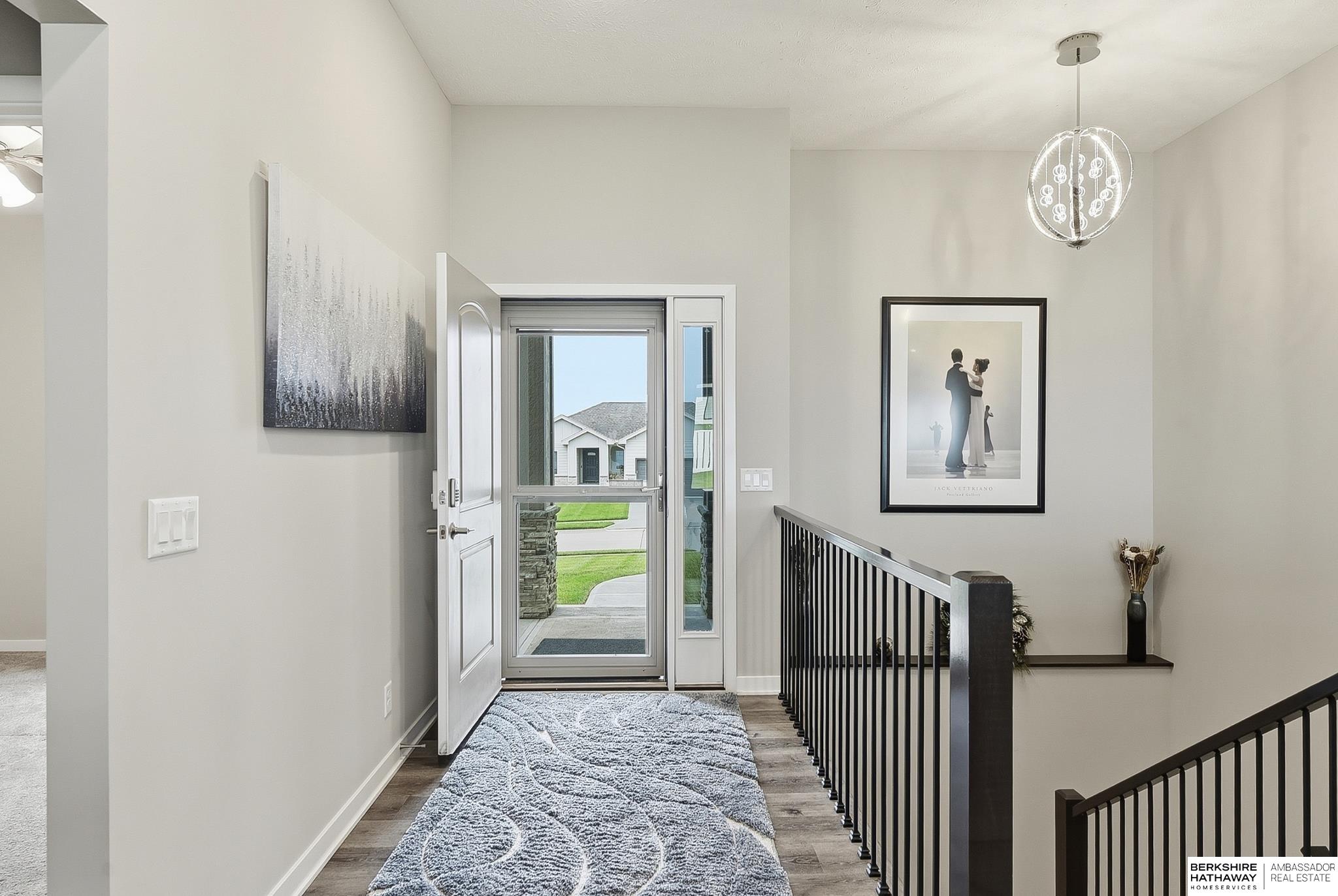 Entry foyer with an open front door, patterned runner, black stair railing, and modern pendant light.