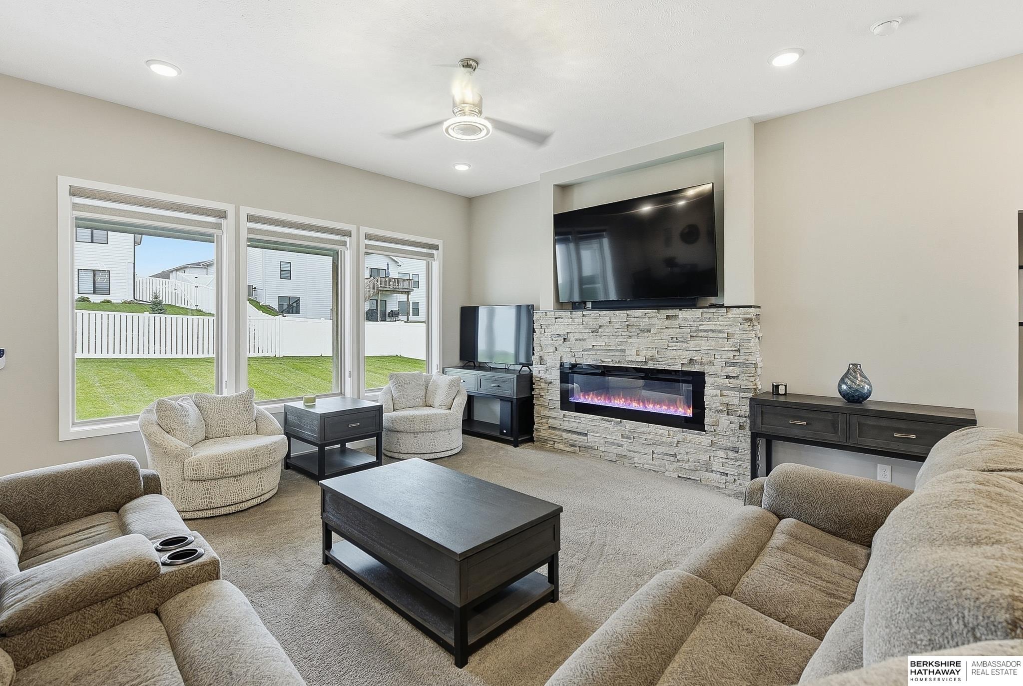 Furnished living room with a stone fireplace, mounted television, and large windows overlooking the backyard.