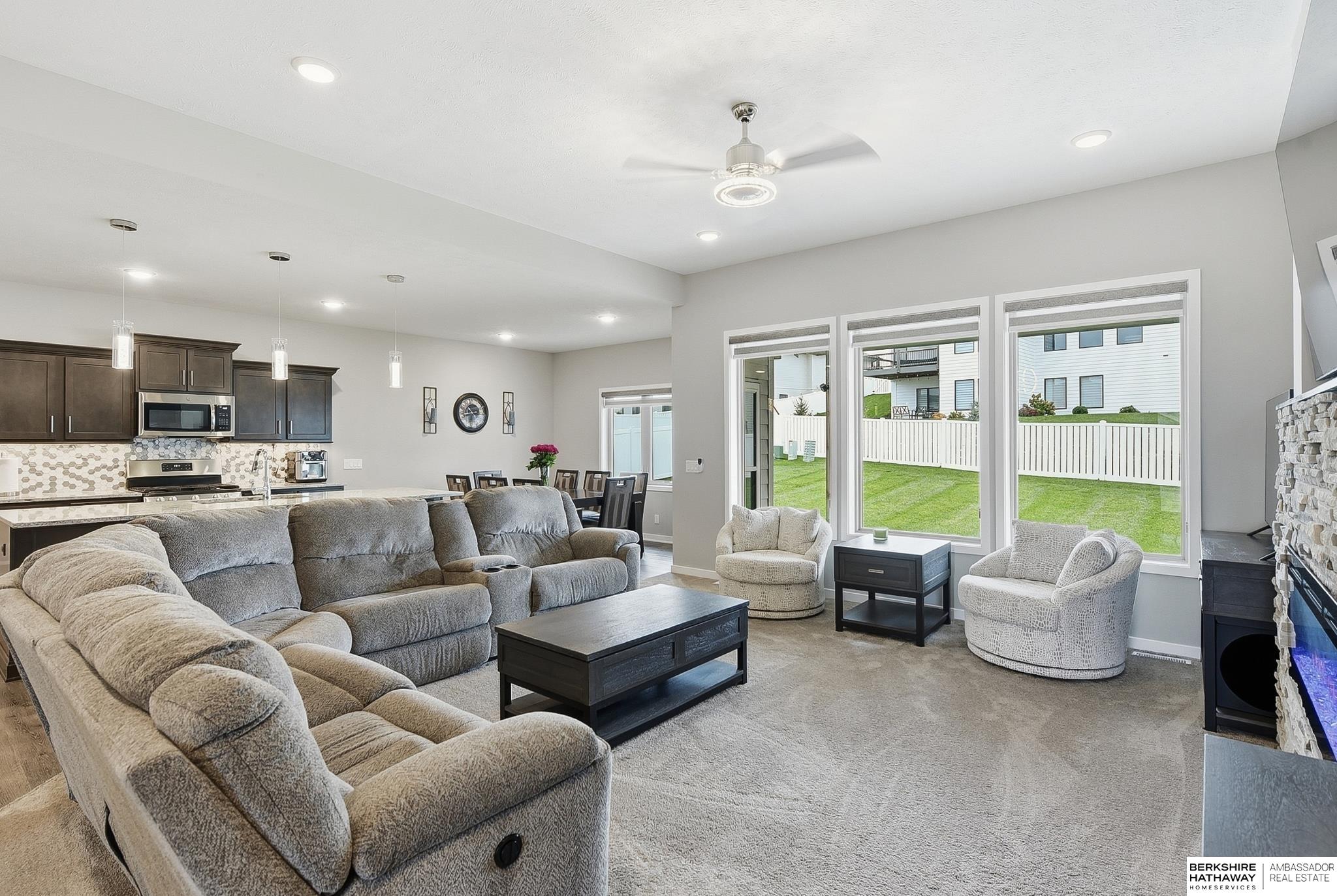 Furnished living room with gray sectional, stone fireplace, and large windows overlooking a fenced lawn.