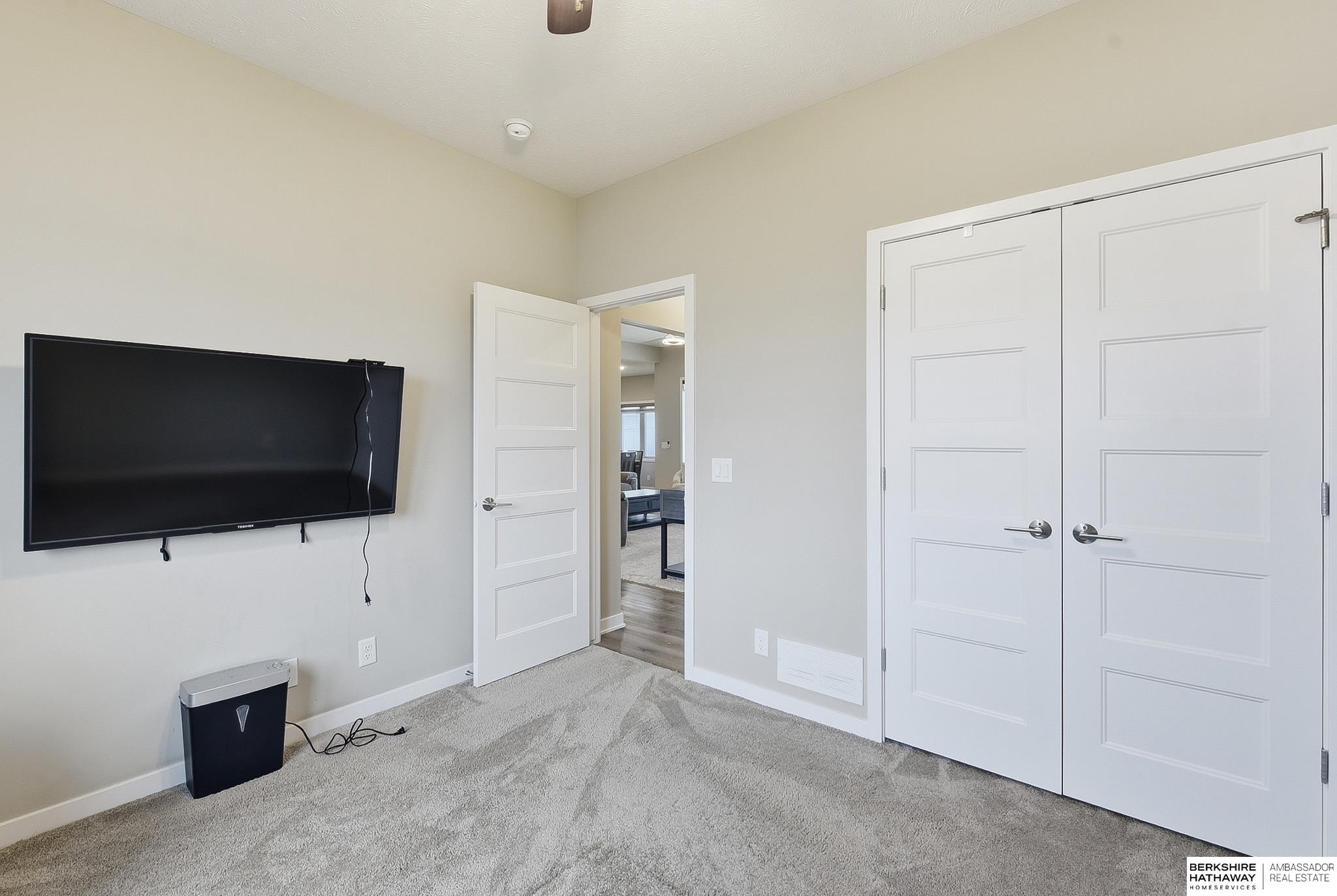 Carpeted bedroom with a wall-mounted television, double closet doors, and an open doorway to a sitting area.