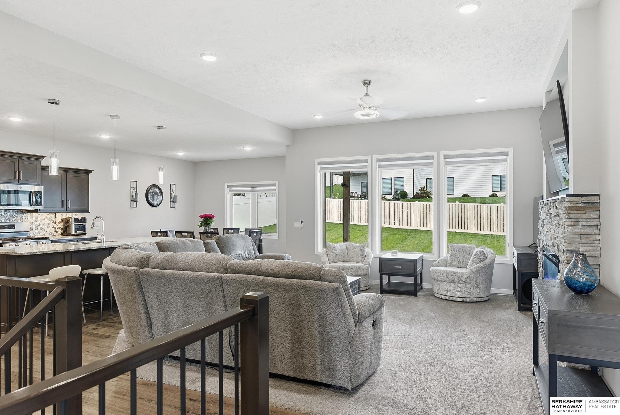 Open living room with gray seating, a stone fireplace, and windows overlooking a fenced yard.