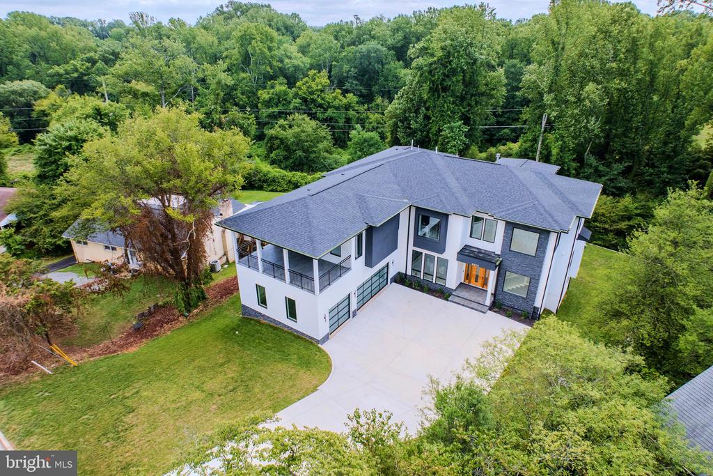 Aerial view of a modern white house with a gray roof, balcony, broad driveway, and wooded surroundings.
