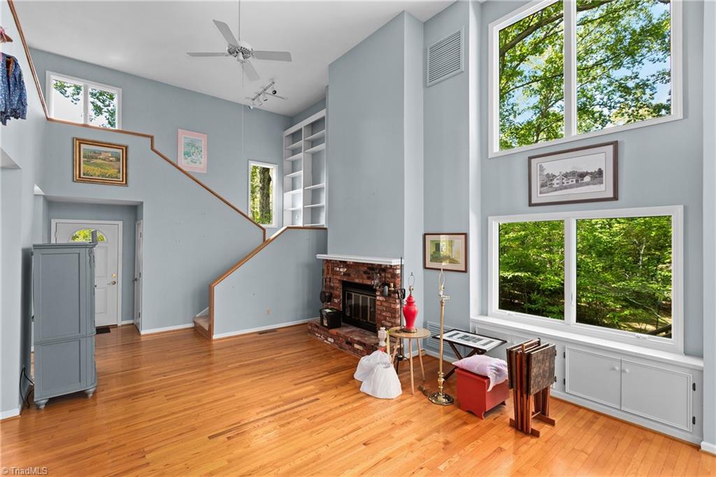 Living room with a brick fireplace, hardwood floors, staircase, and large windows overlooking trees.
