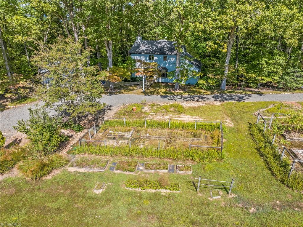 Aerial view of a blue house among trees, with a gravel drive and fenced garden beds.