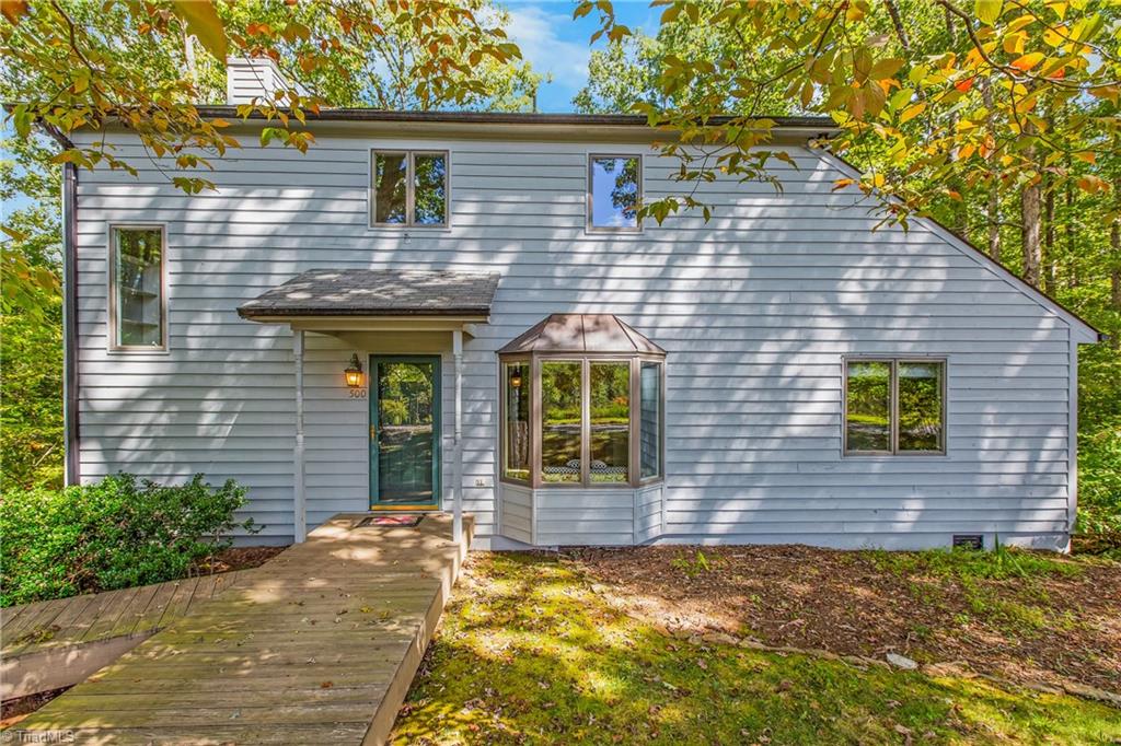 Light-sided home with a projecting bay window and wooden walkway beneath autumn trees.