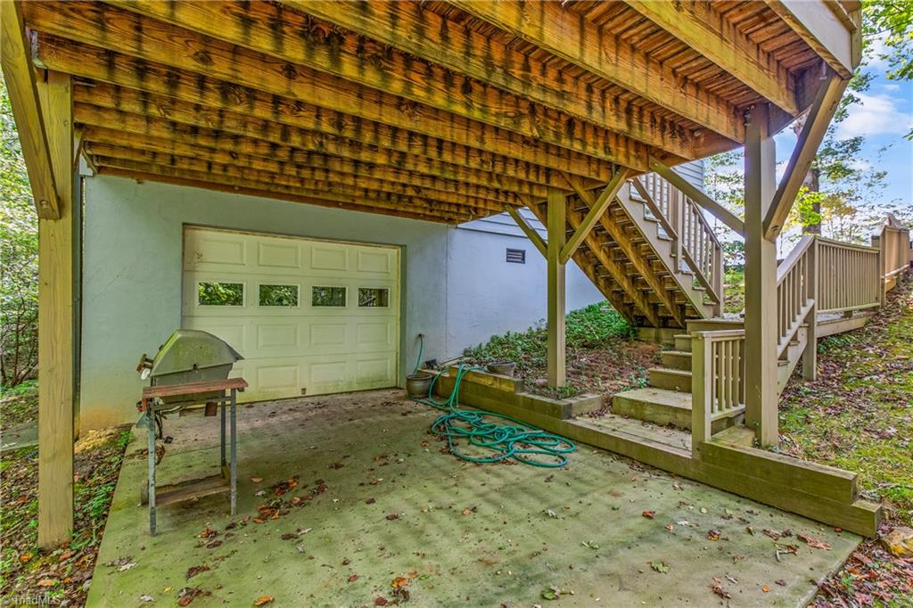 Covered concrete patio beneath a raised wooden deck, with stairs, garage door, and grill.
