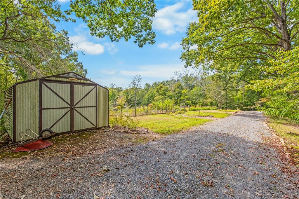 Gravel driveway beside a metal storage shed, opening onto a wooded yard and fenced garden.