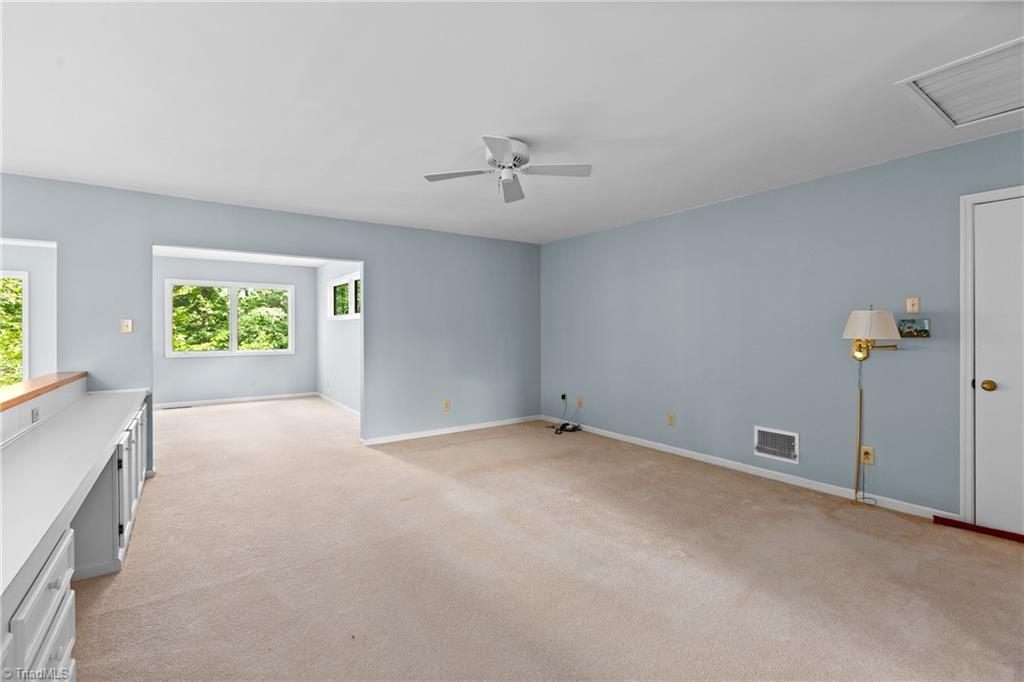 Empty carpeted room with pale blue walls, built-in cabinetry, a ceiling fan, and windows overlooking greenery.