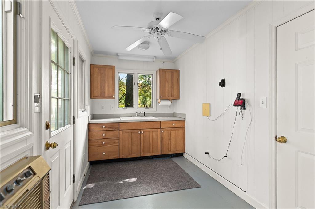 Narrow room with wood cabinets and sink beneath a window, white paneled walls, and a ceiling fan.