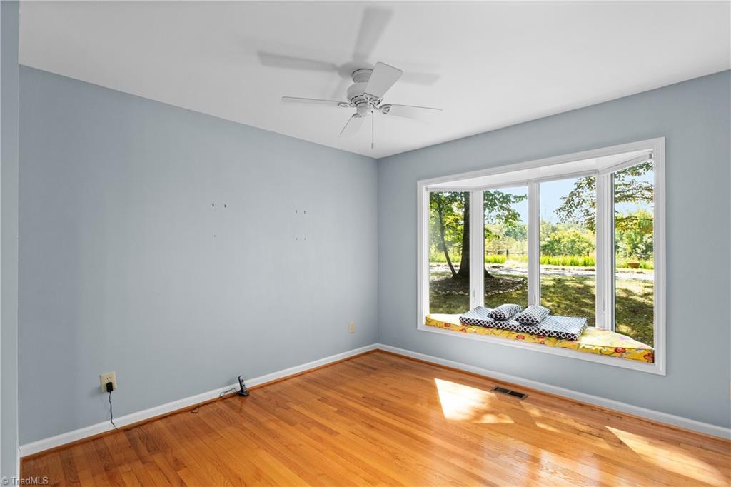 Unfurnished room with hardwood floors, a ceiling fan, and a large window overlooking a leafy yard.