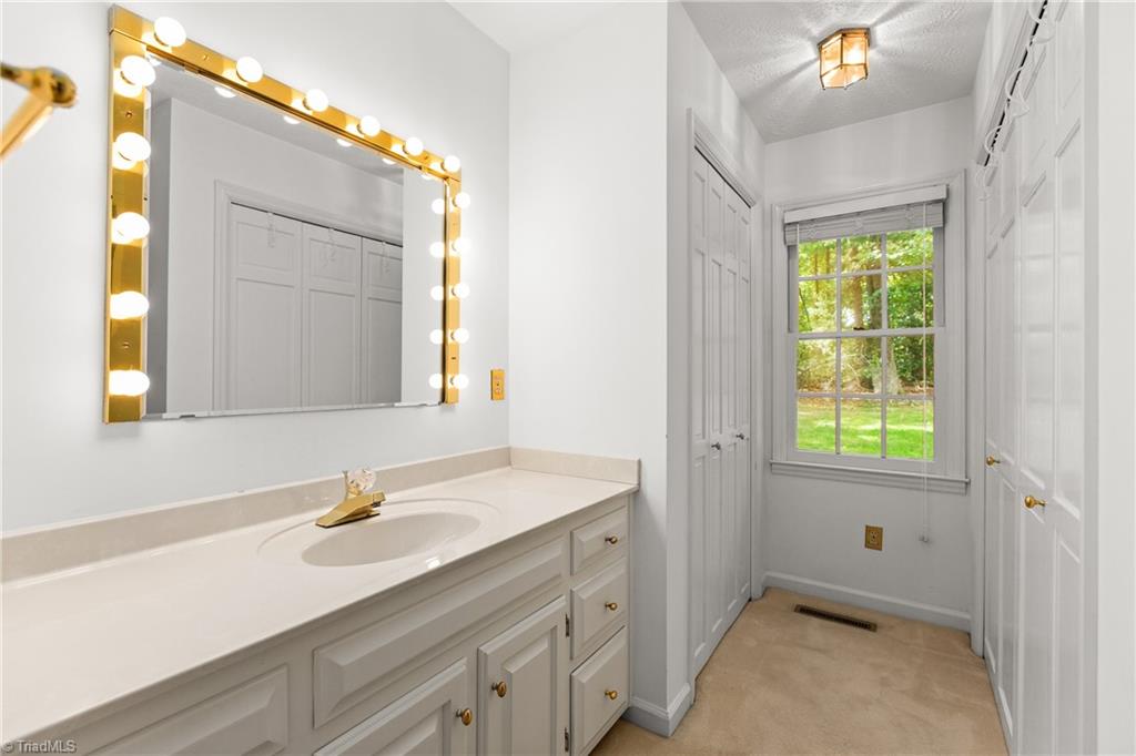 Bathroom with a long white vanity, bulb-lit mirror, and window overlooking trees and lawn.