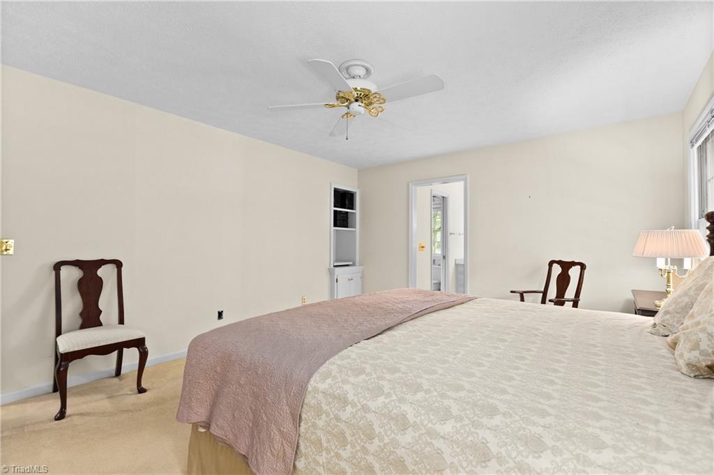 Furnished bedroom with a large patterned bed, mauve throw, wooden chairs, and ceiling fan.