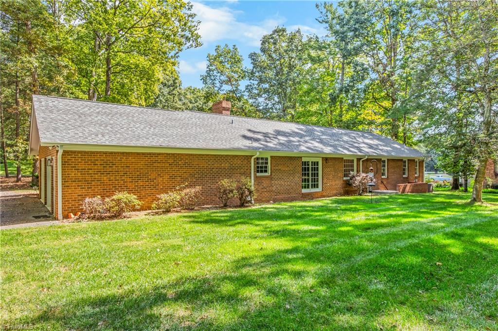 Red-brick home with a long gray roof, brick patio, and mature trees beyond a green lawn.