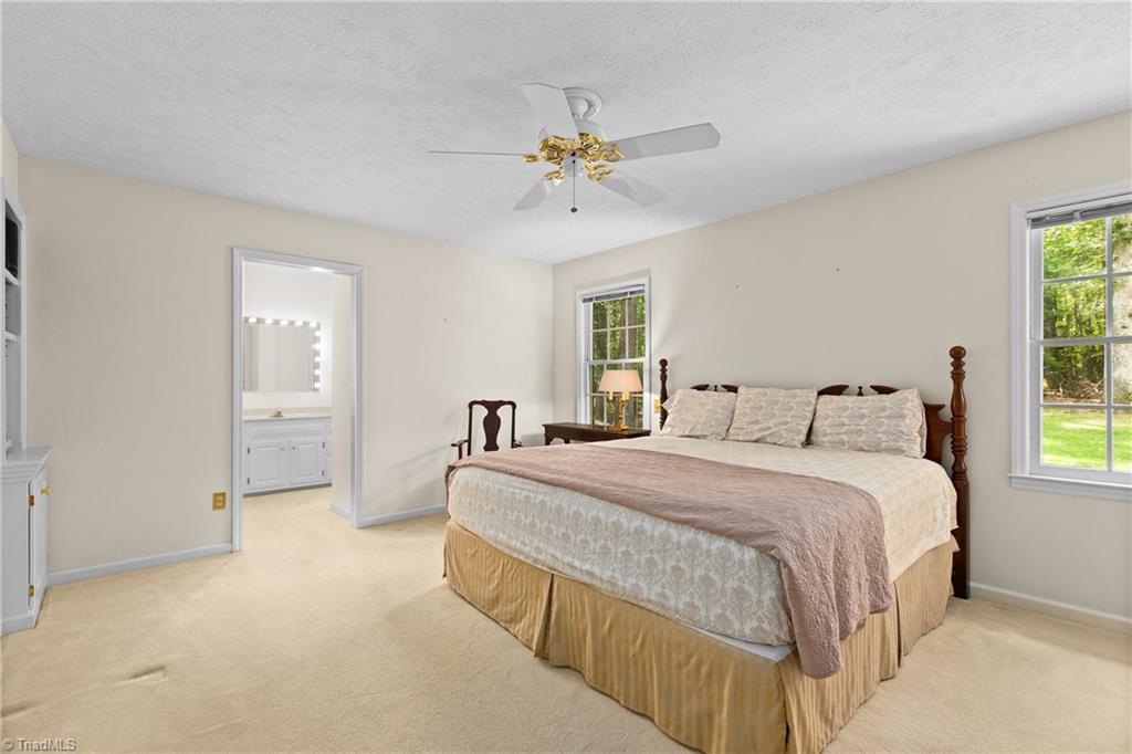 Bedroom with a large bed, beige carpet, ceiling fan, and doorway to an ensuite bathroom.