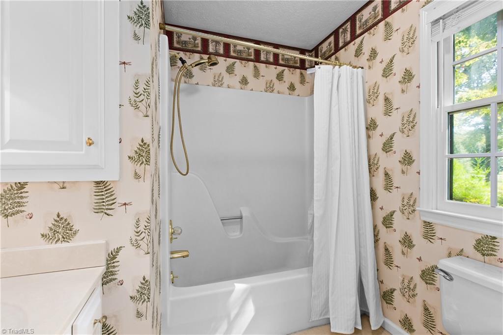 Bathroom with a white tub-shower, brass fixtures, fern-patterned wallpaper, and a bright window.
