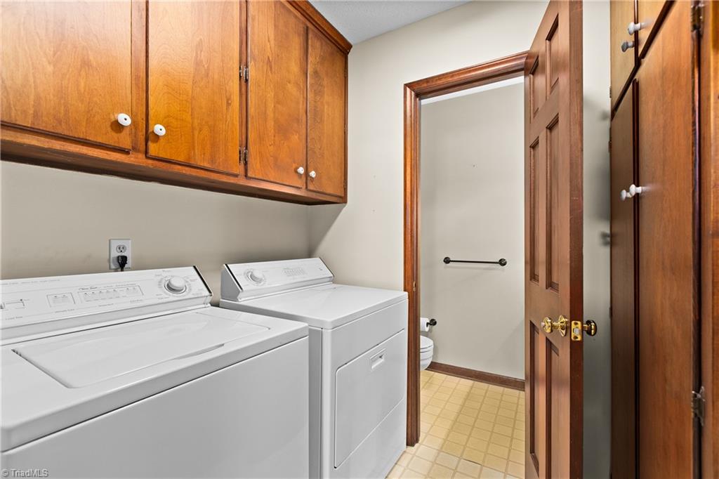 Laundry room with white washer and dryer beneath wood cabinets, opening to a bathroom.