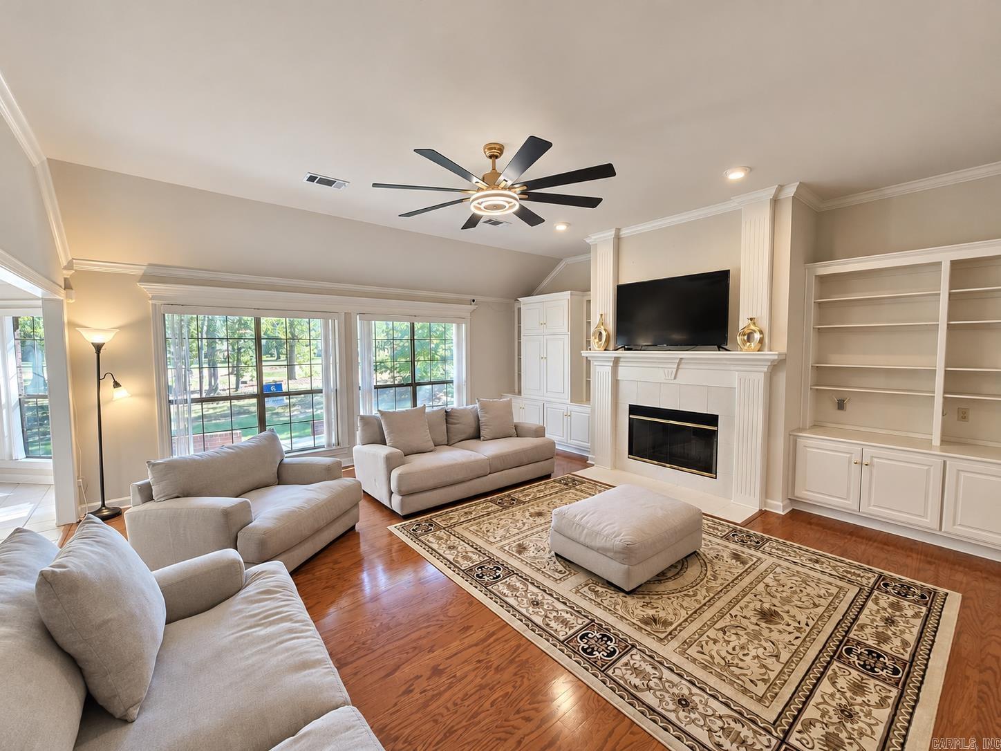 Living room with a white fireplace, built-in shelving, hardwood floors and large windows overlooking greenery.