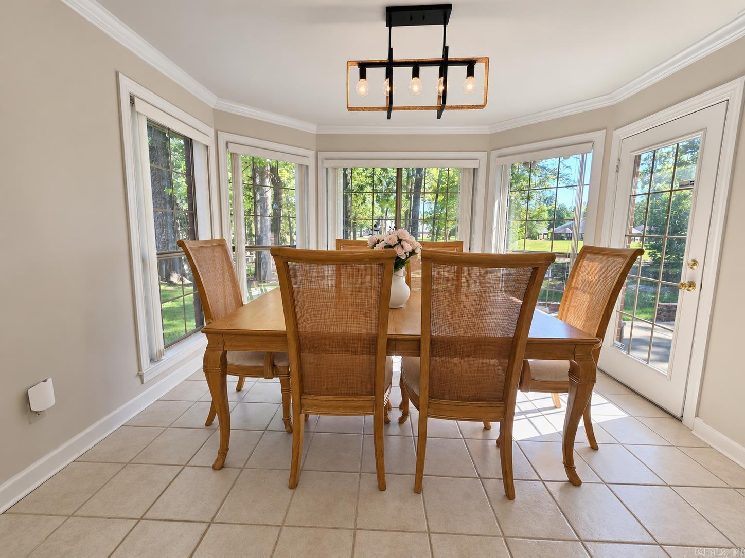 Dining room with a wooden table and cane-back chairs surrounded by tall windows overlooking a leafy yard.