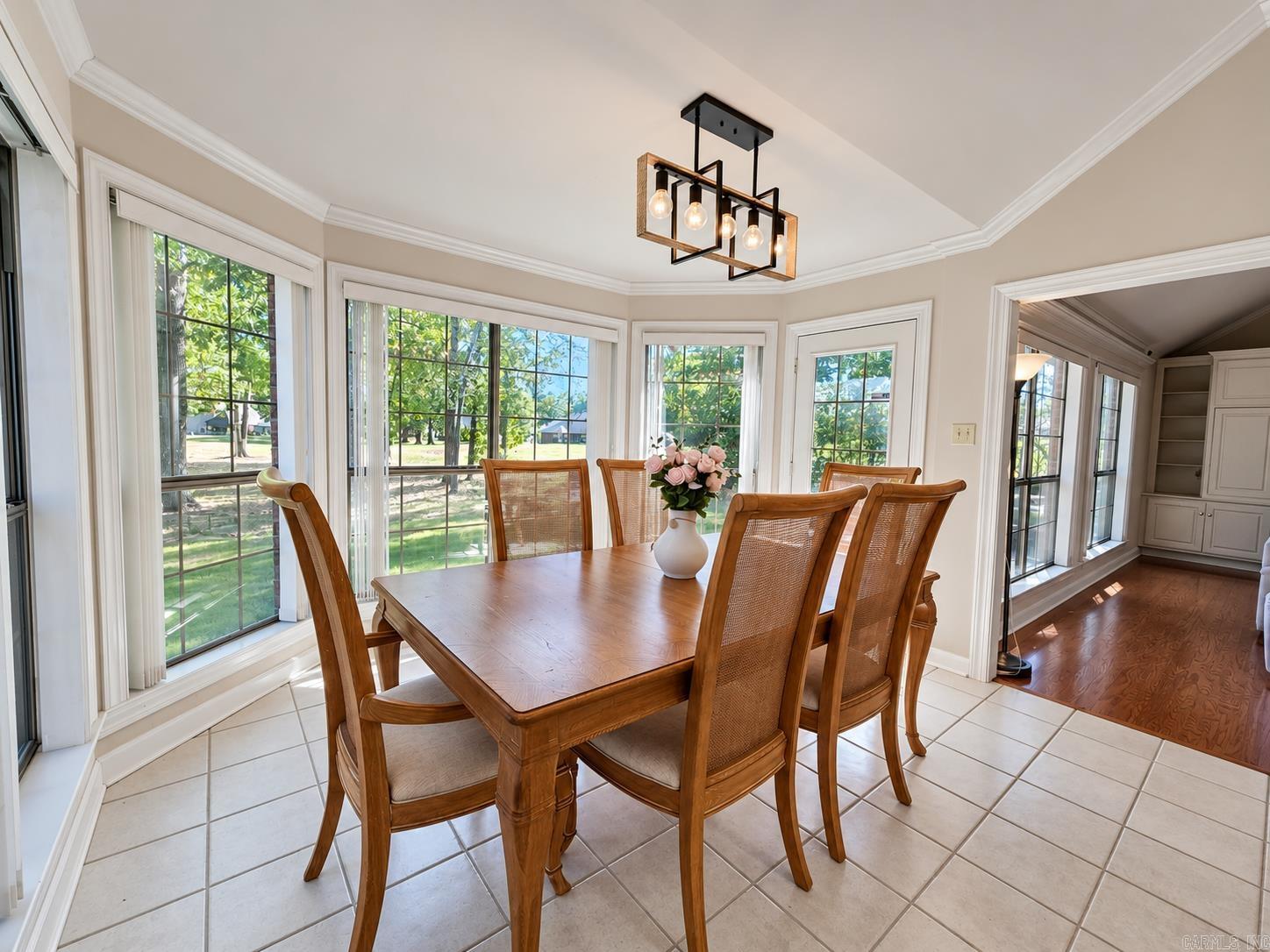 Dining room with a wooden table and chairs beside large windows overlooking a leafy yard.