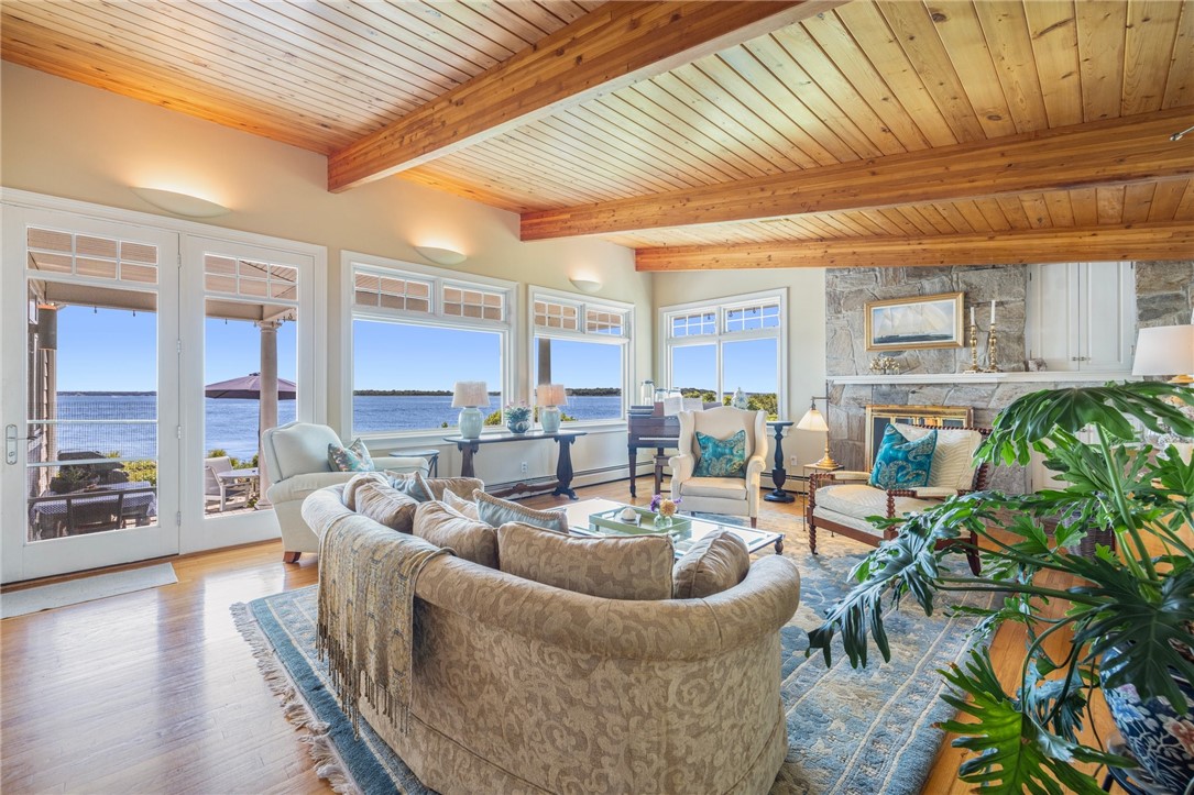 Living room with exposed wood-beam ceiling, stone fireplace, and water views through large windows.