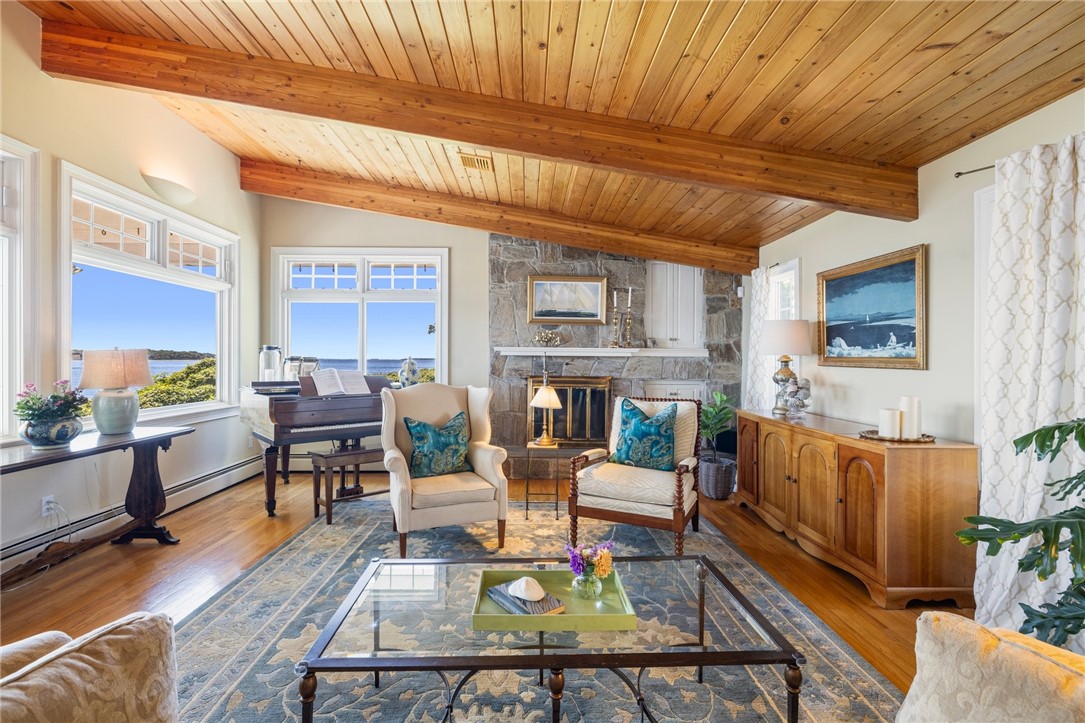 Living room with vaulted wood ceiling, stone fireplace, grand piano, and water views through large windows.