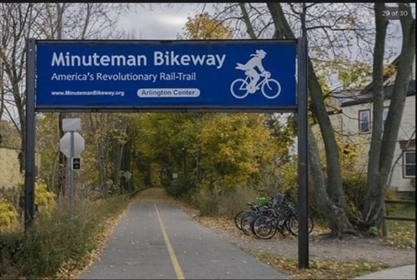 Minuteman Bikeway path beneath its Arlington Center sign, with autumn foliage and bicycles nearby.