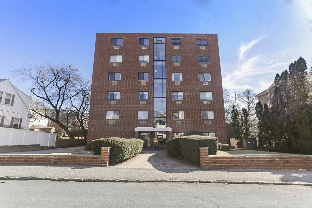 Red-brick apartment building with a central glass bay and hedges lining the entrance walkway.