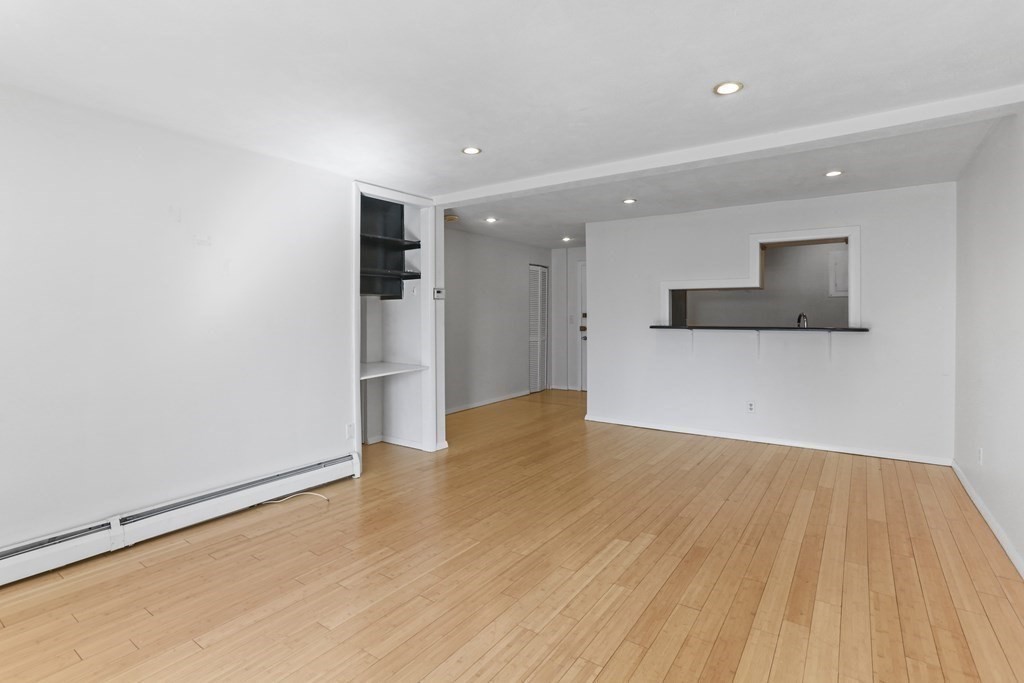 Empty living room with hardwood floors, built-in shelving, and a pass-through to the kitchen.