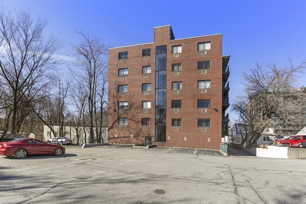 Red brick apartment building with a central glass window section, side balconies, and a parking lot.