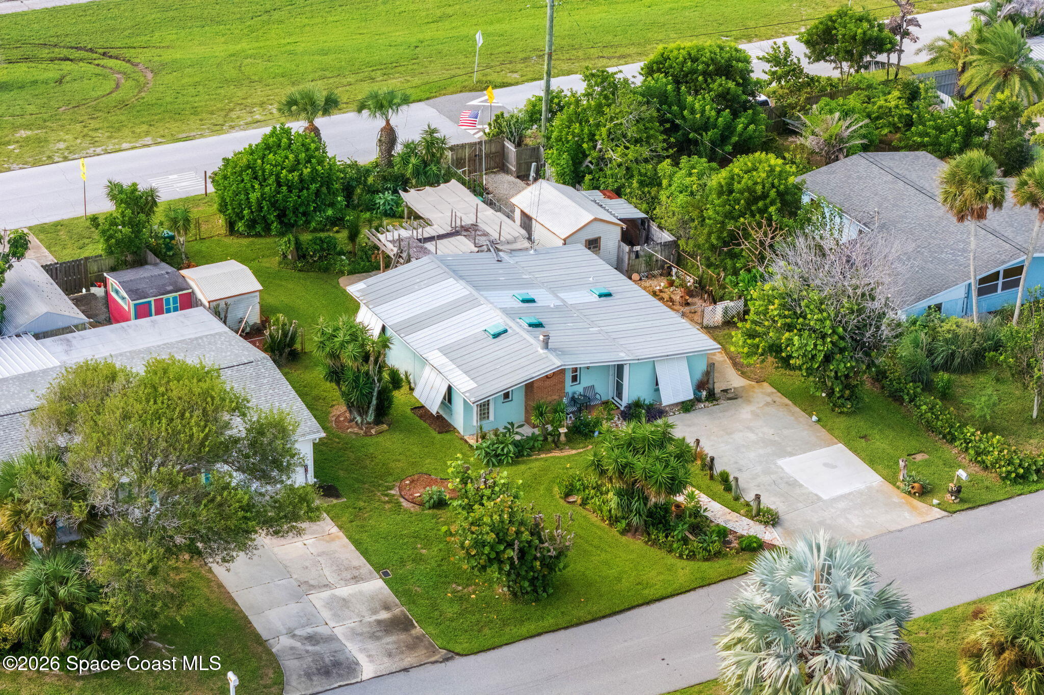 Aerial view of a light-blue home with a broad metal roof, lush landscaping, and a large driveway.